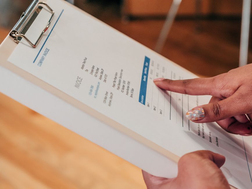 Imagem de close-up de uma pessoa apontando para um documento em um clipboard, dentro de casa.