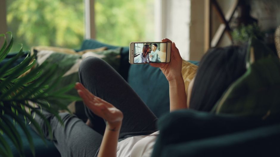 Mulher relaxando em um sofá, participando de uma chamada de vídeo em seu smartphone, dentro de casa.